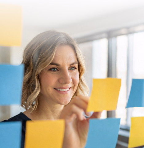 A diverse group of professionals in business attire converse animatedly in a modern office lobby. They wear blue lanyards and appear engaged and smiling.